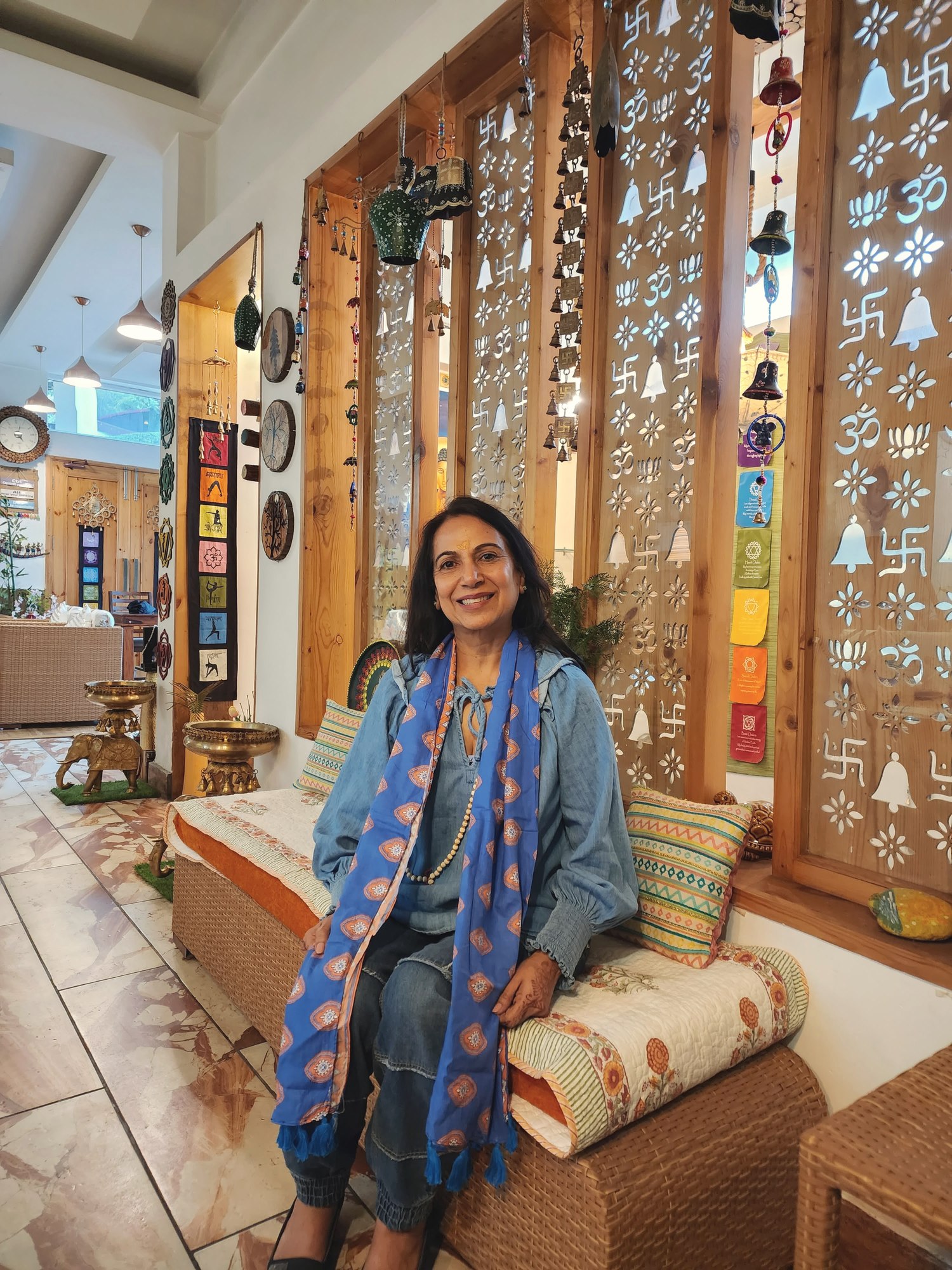 Neetu smiling warmly, seated in a calming space decorated with bells and Om symbols