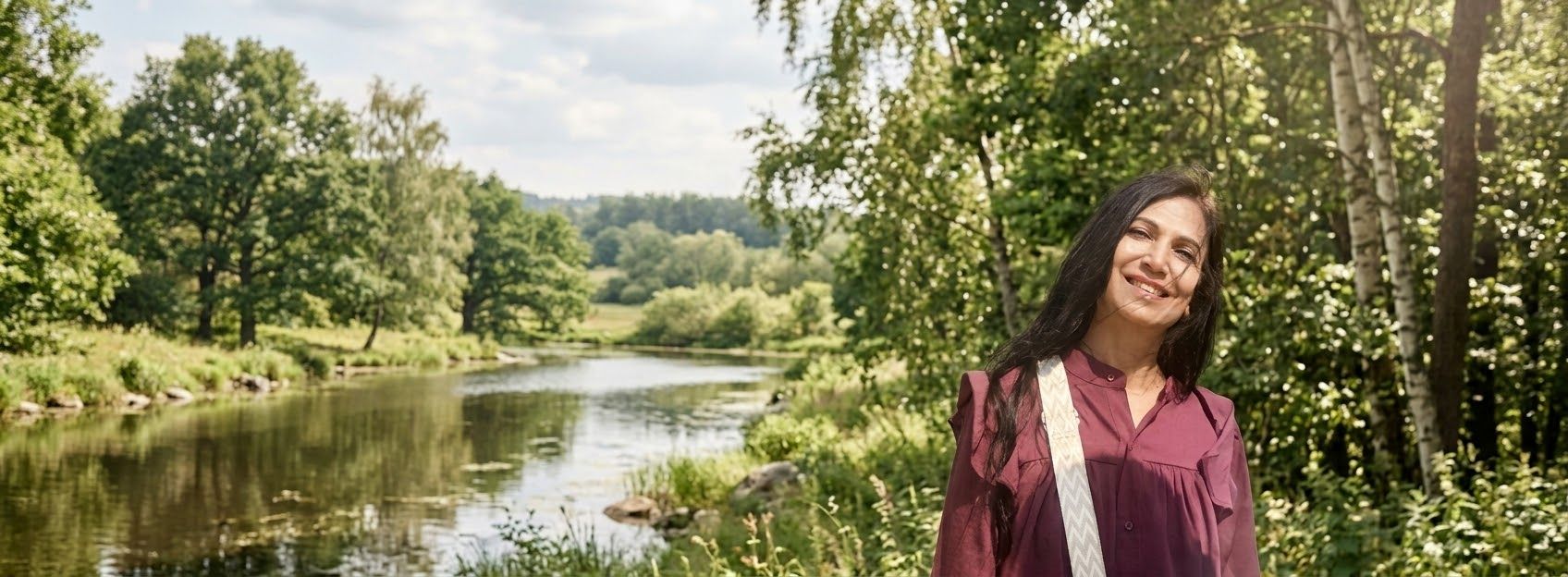 Neetu smiling warmly beside a winding river, surrounded by trees